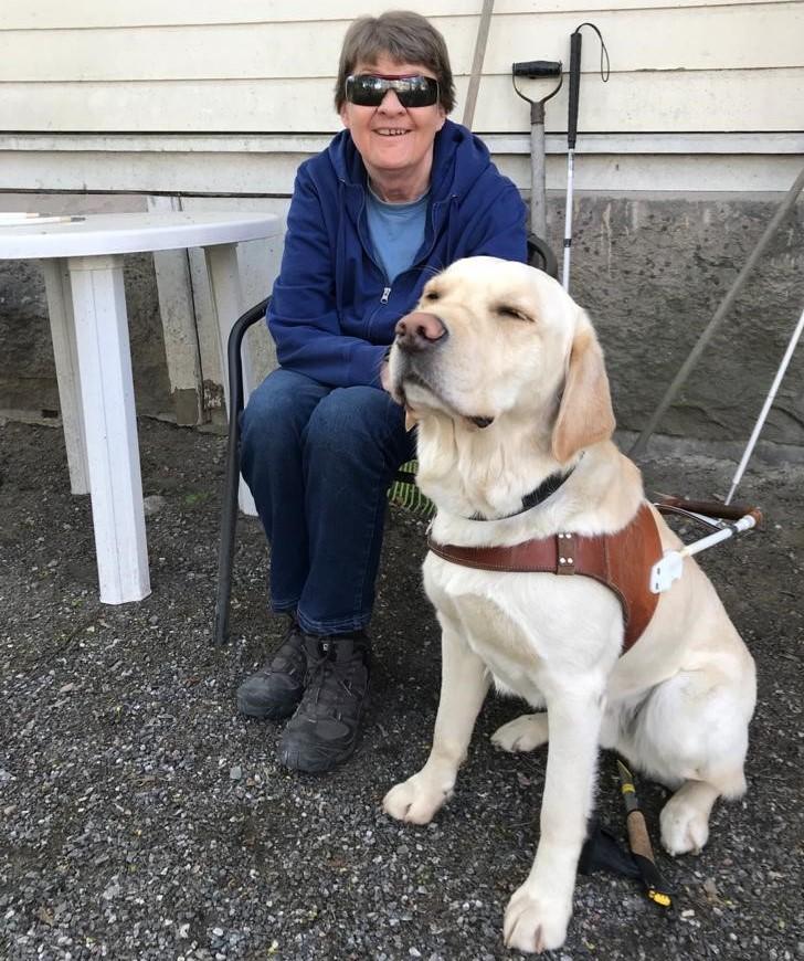 Marita istuu tuolilla ulkona vierellään opaskoira, vaalea labradorinnoutaja. Taustalla vaalea talon seinä.